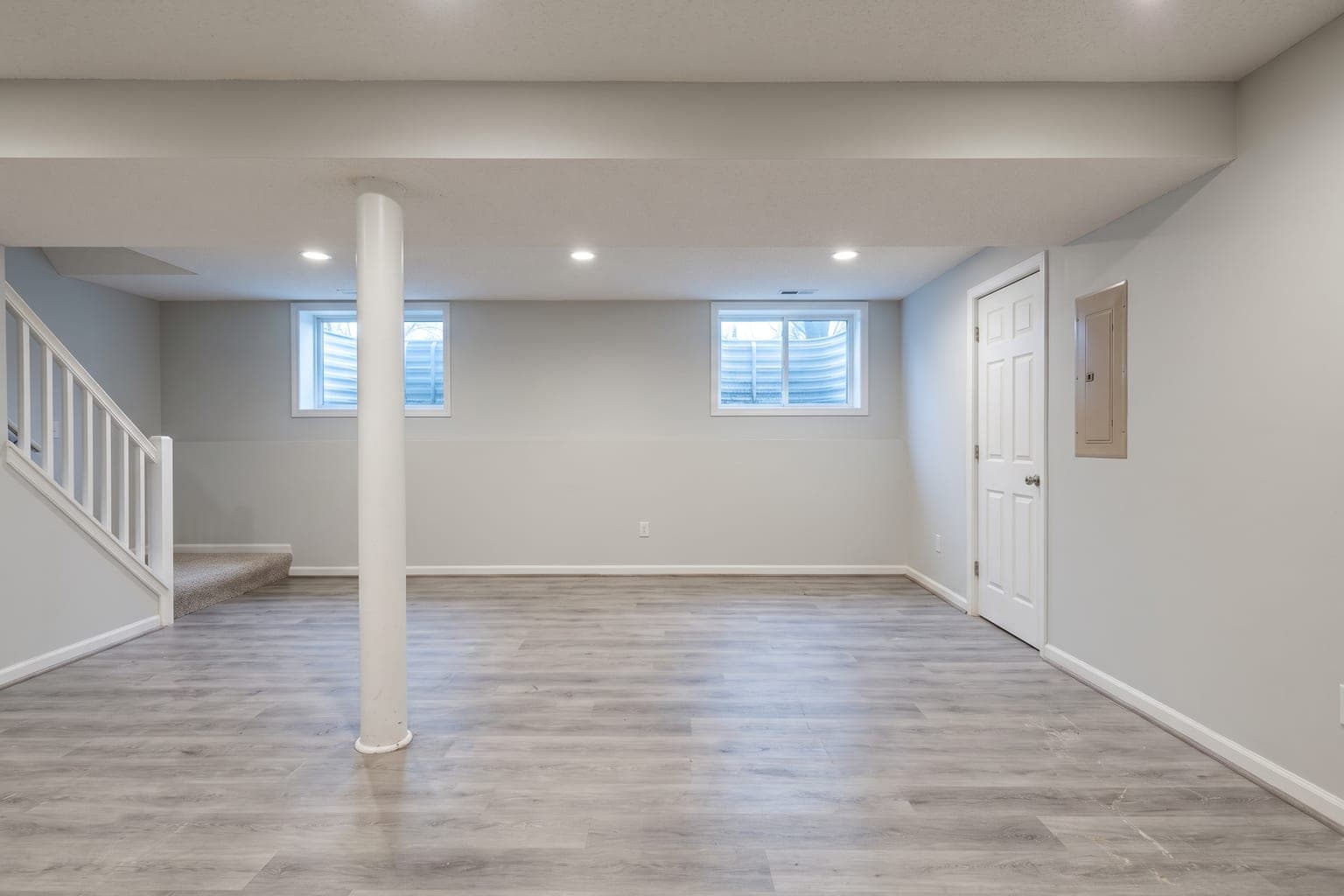 Empty finished basement before virtual staging - gray vinyl plank floor, low ceiling, egress windows, support column