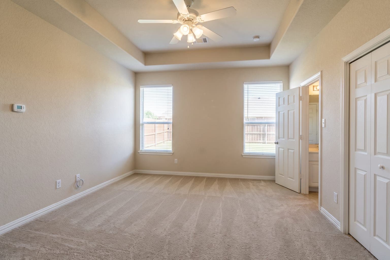 Empty carpeted primary bedroom before virtual staging - beige walls, tray ceiling, two windows, closet doors