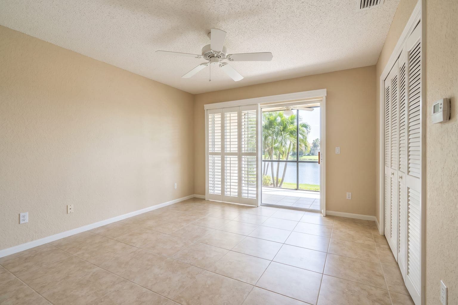 Empty Florida condo bedroom before virtual staging - beige tile, plantation shutters, sliding door to a lanai with lake view