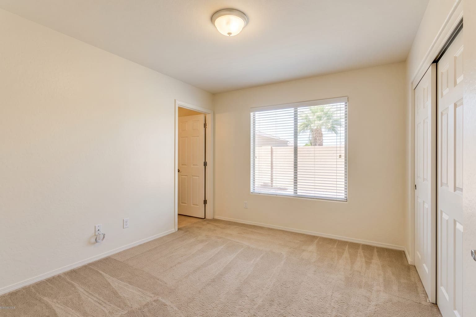 Empty carpeted spare bedroom before virtual staging - off-white walls, window with blinds, bifold closet doors