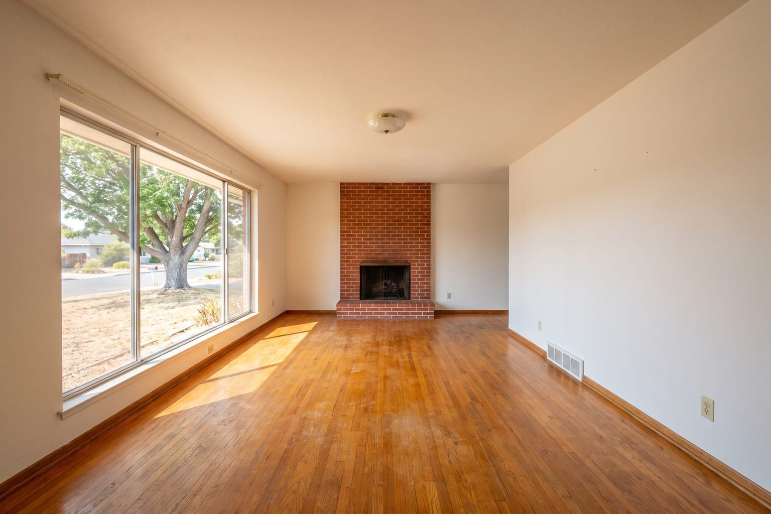 Empty 1960s ranch living room before virtual staging - oak floors, brick fireplace, large picture window